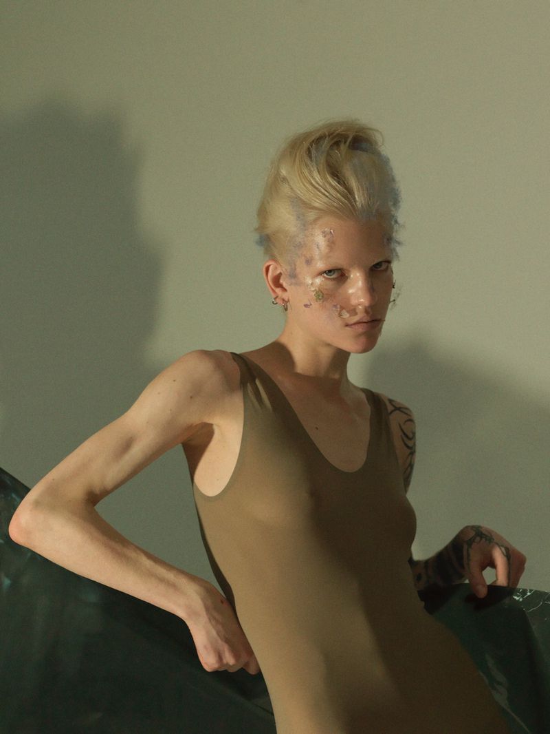 Portrait of a woman with blond hair and body glitter, wearing a brown leotard, posed in front of a plain light green background.