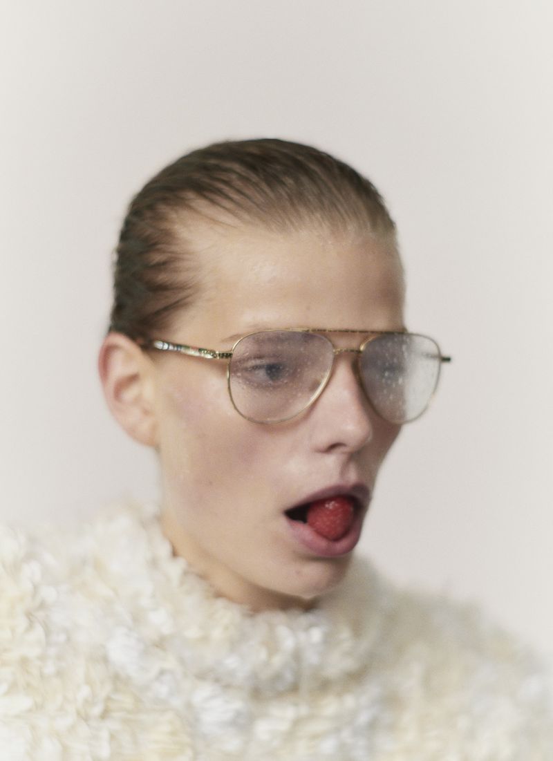 Close-up portrait of a young woman wearing aviator glasses with water droplets, holding a cherry on her tongue, with a soft-focus background.
