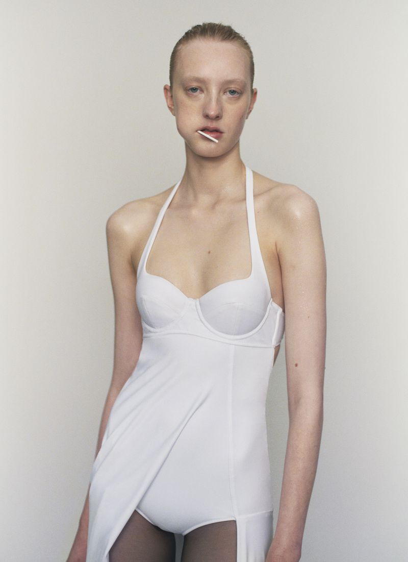 A model poses in a white outfit against a plain backdrop, featuring bright lighting and a minimalist aesthetic. A lollipop is in the model's mouth.