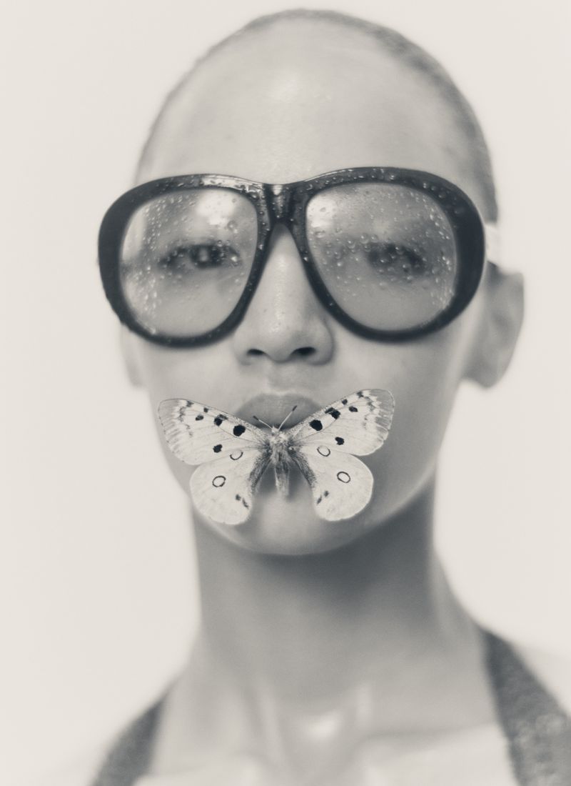 Close-up portrait of model Ebba Bostrom wearing oversized dark goggles with misted lenses and a butterfly resting on her lips.