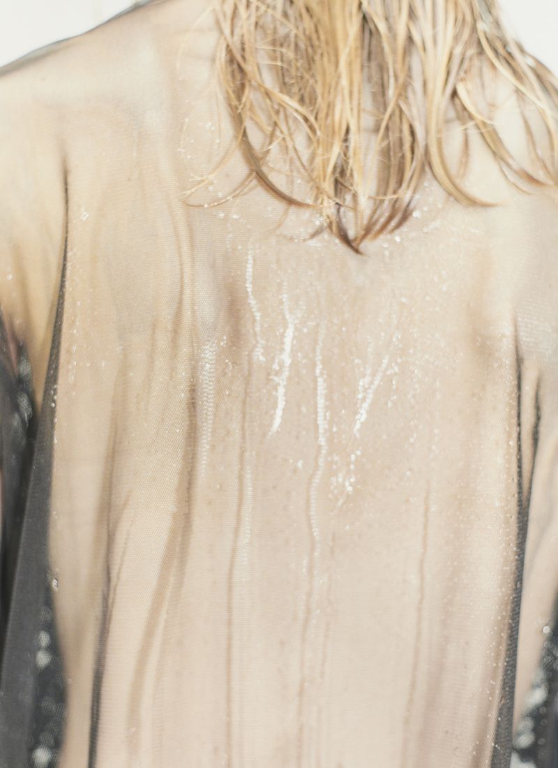 Close-up rear view of model with wet blonde hair wearing translucent sheer fabric with water streaks.