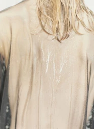 Close-up rear view of model with wet blonde hair wearing translucent sheer fabric with water streaks.