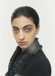 Close-up portrait of a woman with slicked-back dark hair wearing a black ruffled jacket against a plain white background