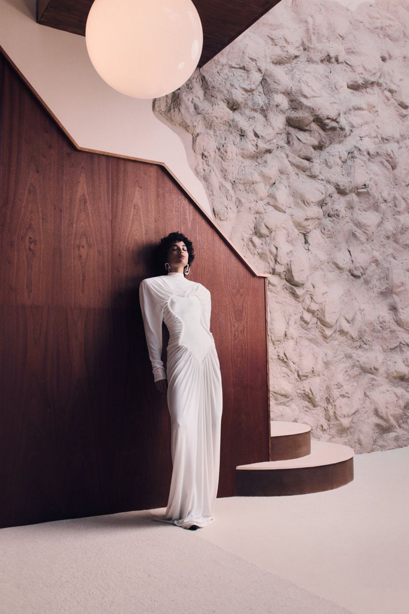 A woman in a white dress poses in front of a brown and white wall with a large round light fixture in an indoor architectural setting.