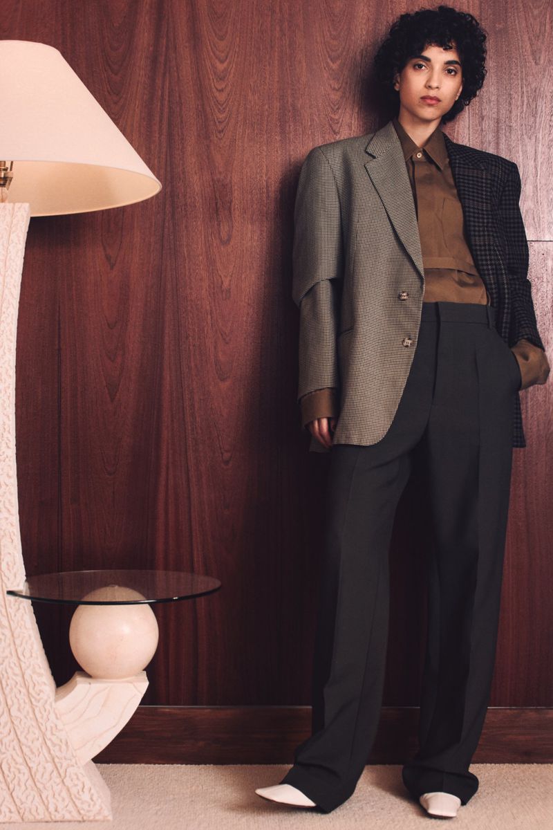 A woman with curly hair poses indoors wearing a brown and grey blazer, brown shirt, black pants, and white shoes against a brown wall.