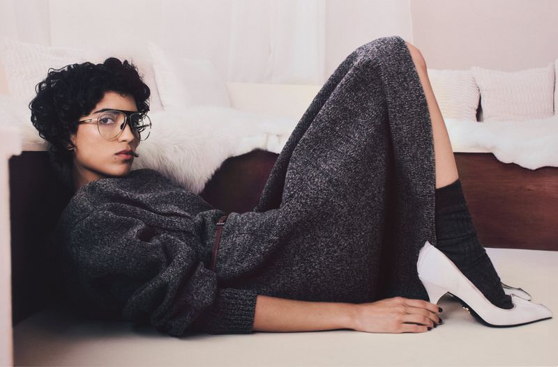Indoor fashion shot of an adult female model in a gray sweater dress, glasses, black socks, and white heels, posing on the floor.