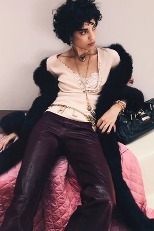 Model Santana reclining in burgundy leather trousers, a cream lace-trimmed top, black fur stole, and layered gold jewellery.