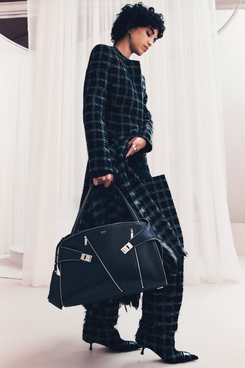 Model in a dark plaid coat and matching boots holding a large black structured handbag against draped white fabric.
