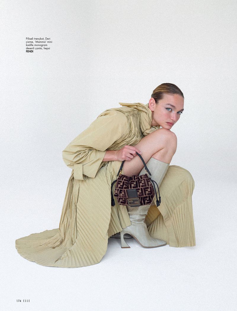 A woman poses in a tan coat, dress, and boots, holding a brown monogrammed Fendi purse, set against a stark white background.
