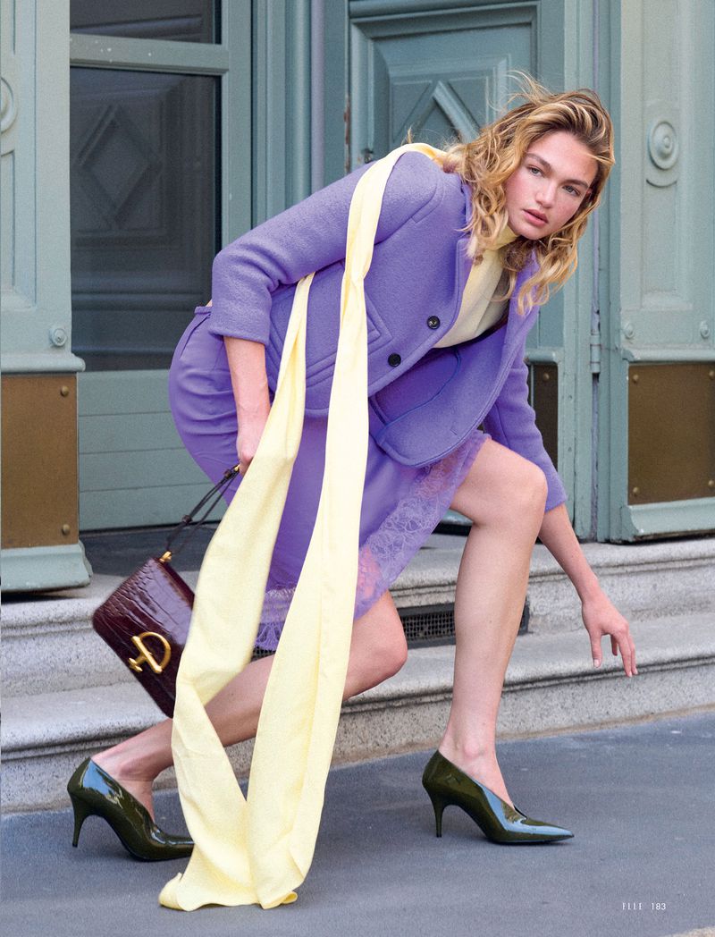 Woman poses on steps in a purple skirt suit with a yellow scarf, handbag, and green high heels, creating a fashion editorial shot.