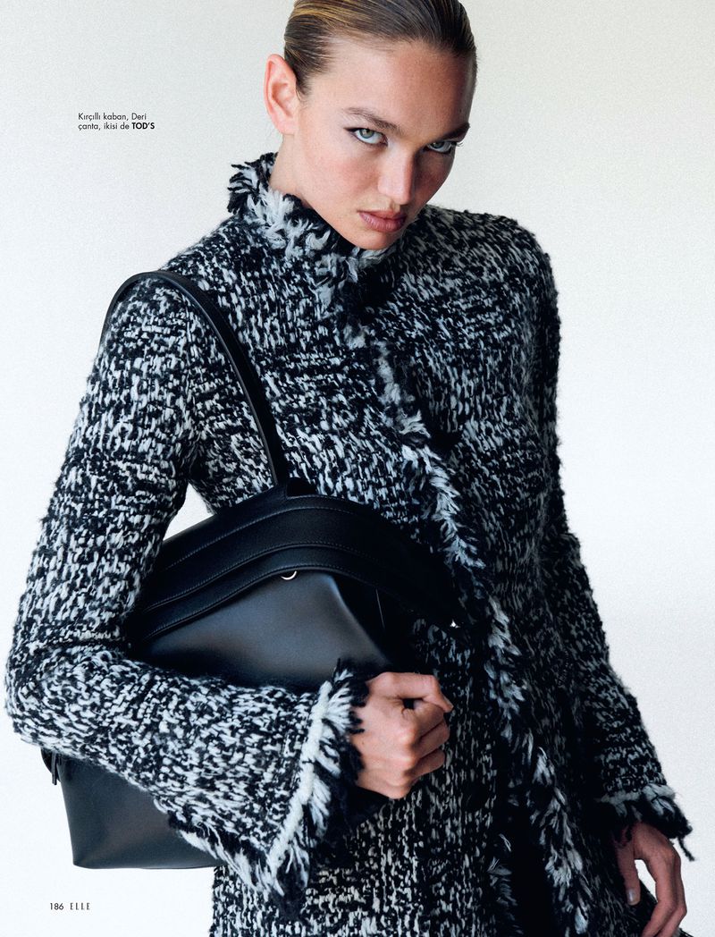 A woman models a black and white textured coat with a black leather handbag in a studio, against a white backdrop. Fashion editorial.