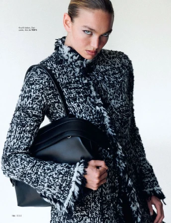 A woman models a black and white textured coat with a black leather handbag in a studio, against a white backdrop. Fashion editorial.