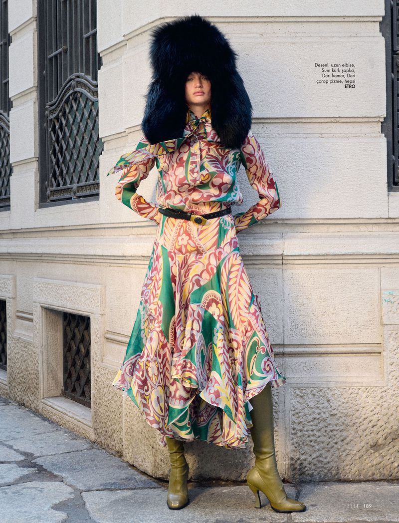 Fashion shot of a woman in a patterned dress and high-heeled boots, wearing a large fur hat and standing on a city street.