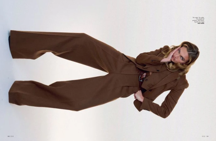 Fashion editorial shot of a blonde adult woman wearing a brown suit and posing against a white backdrop.