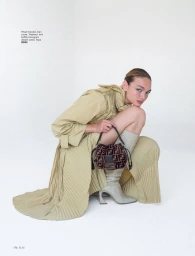 Camille Opp crouching in a pleated beige coat with high-heeled boots and a patterned handbag against a white studio background.