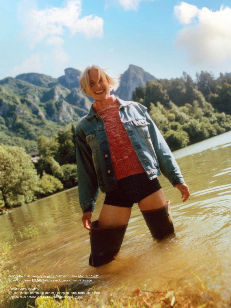 Woman standing in shallow water with tall rubber boots, wearing a denim jacket, with mountains and a cloudy blue sky in the background.