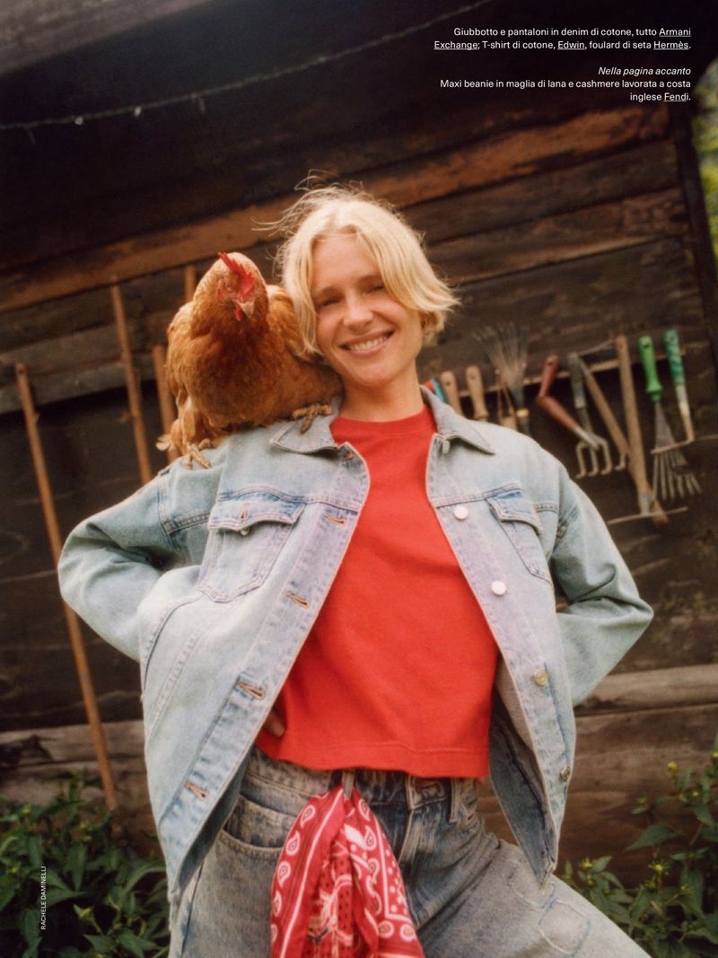 A smiling blonde woman poses outdoors with a chicken perched on her shoulder, wearing a denim jacket and jeans with a red top.