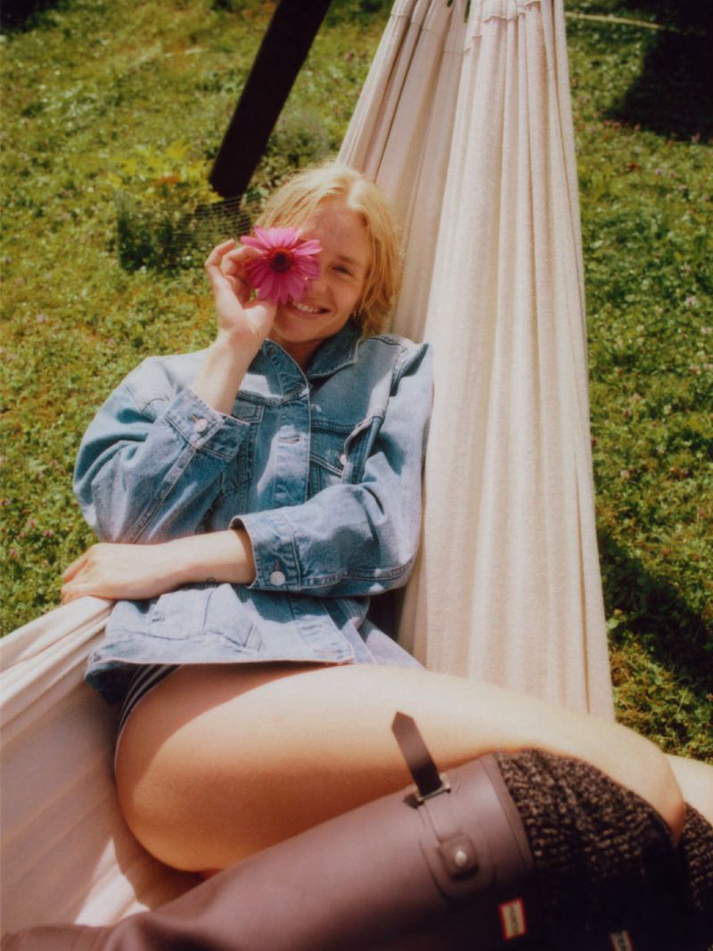 A smiling woman in a denim jacket and black boots relaxes in a hammock, holding a pink flower to her eye in a sunlit outdoor setting.
