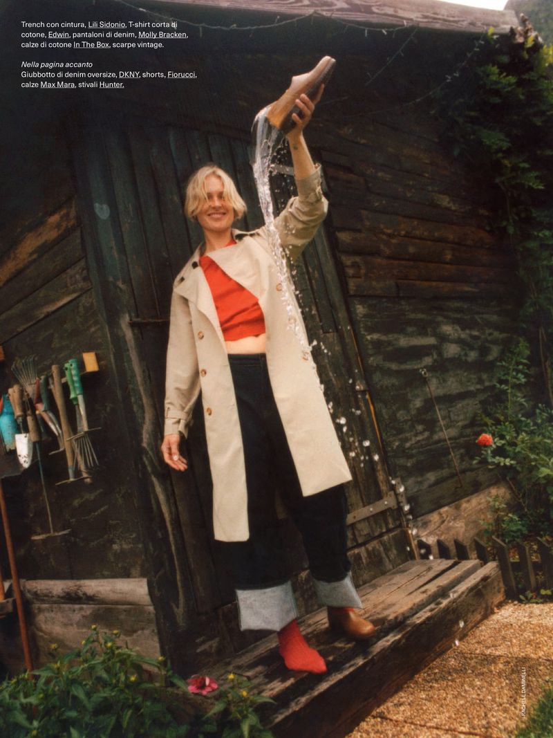 A blonde woman in a trench coat stands smiling outside a rustic wooden shack, playfully pouring water from a wooden clog.