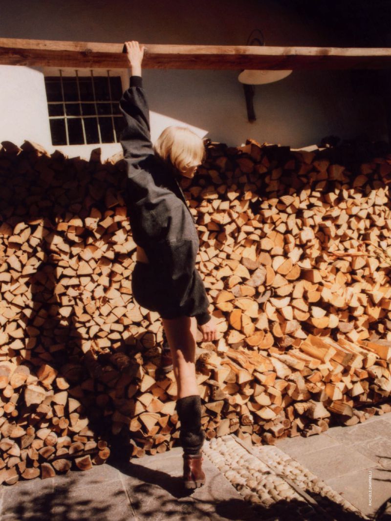 A slender adult woman with short blond hair is leaning against a stack of logs, wearing a dark jacket and socks in an outdoor setting.