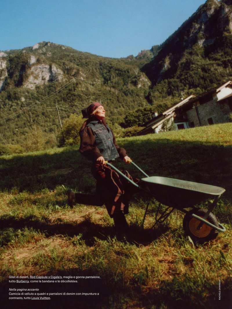 An elderly woman pushes a wheelbarrow across a grassy field, backed by a mountainside and a bright blue sky for a fashion editorial.