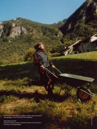 An elderly woman pushes a wheelbarrow across a grassy field, backed by a mountainside and a bright blue sky for a fashion editorial.