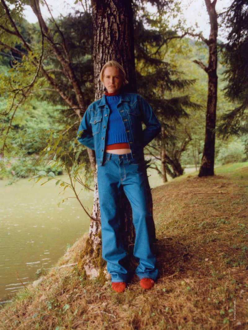 Model Steffi Cook leaning against a tree trunk wearing a denim jacket, blue knit sweater, and jeans in an outdoor forest setting.