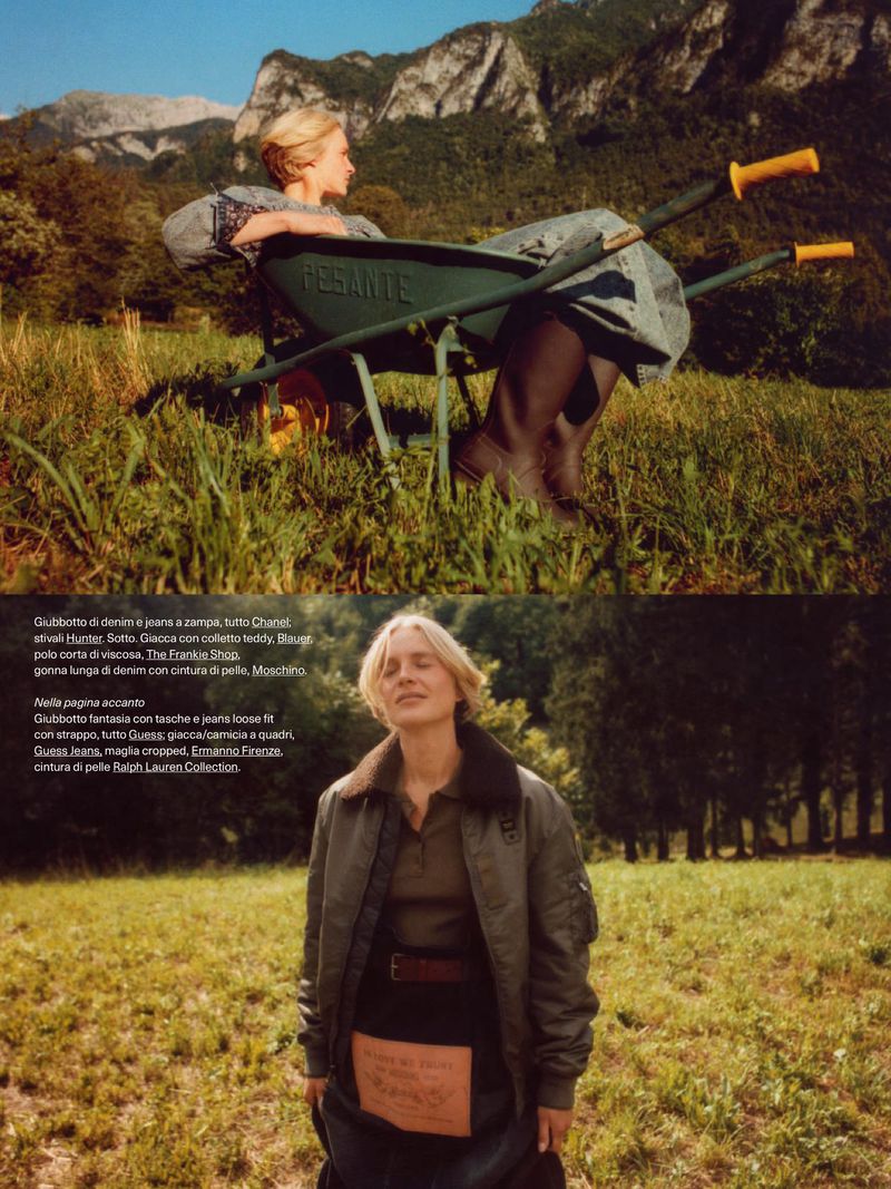 A woman is reclining in a green wheelbarrow in a field with a mountain backdrop. Another woman stands in a field wearing a jacket.