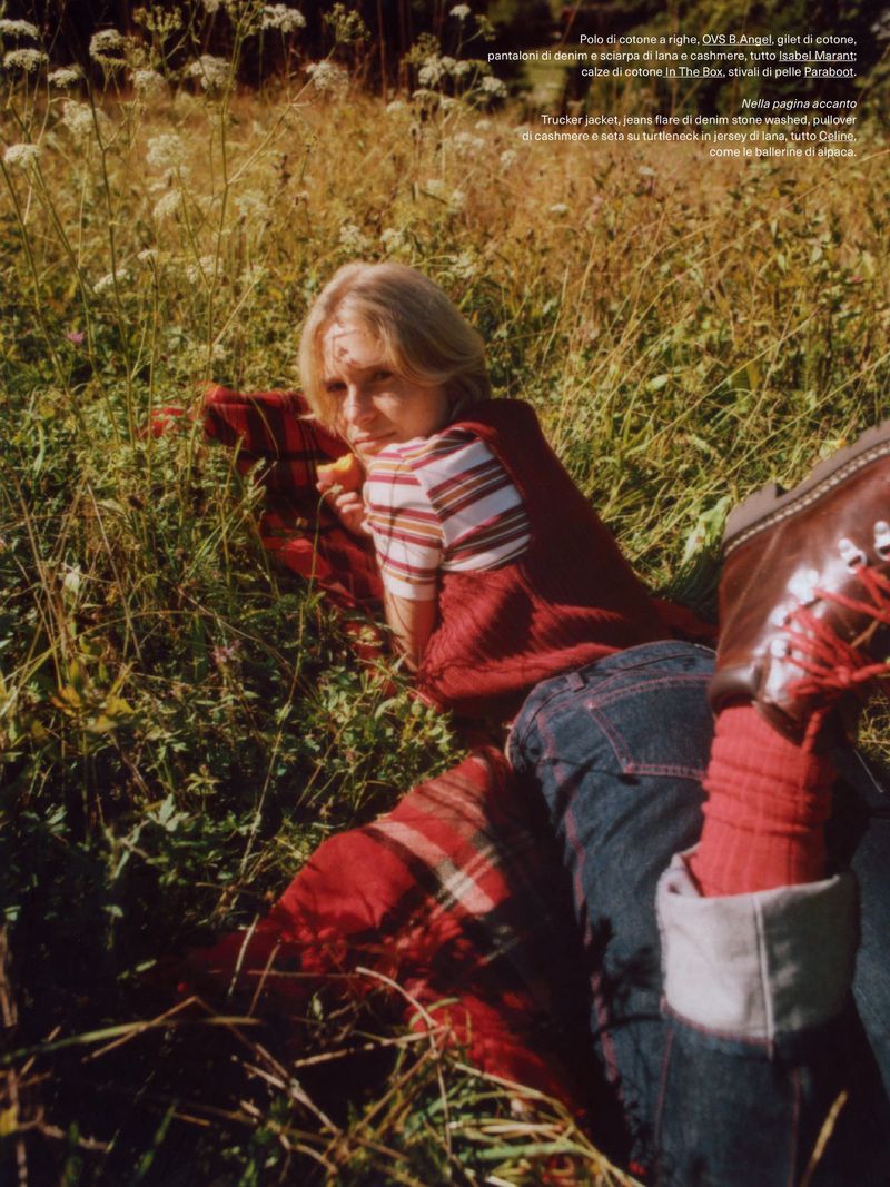 Blonde woman in striped shirt and denim jeans lies on red plaid blanket in grassy field with wildflowers, wearing red socks and leather boots.