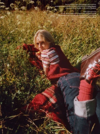 Blonde woman in striped shirt and denim jeans lies on red plaid blanket in grassy field with wildflowers, wearing red socks and leather boots.