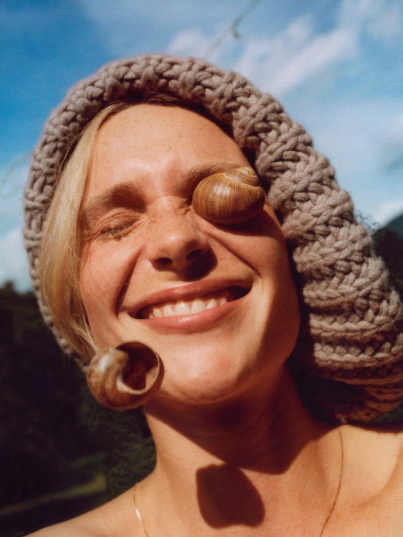 Smiling woman in a knit hat poses with snails on her face in an outdoor setting with blue skies and trees in the background.