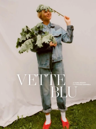 A woman in a denim jacket and jeans stands in front of a white sheet, wearing a leaf crown and holding a leafy branch in her hand.