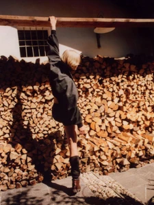 Model Steffi Cook hanging from a wooden beam wearing a dark denim jacket in front of a tall stack of chopped firewood.