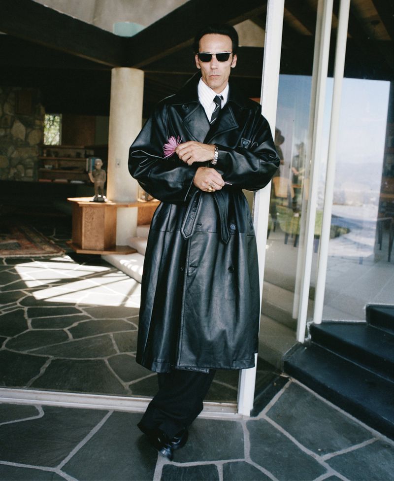 Stylish man in a black leather trench coat poses indoors with a distinctive stone floor and a bright window in the background.
