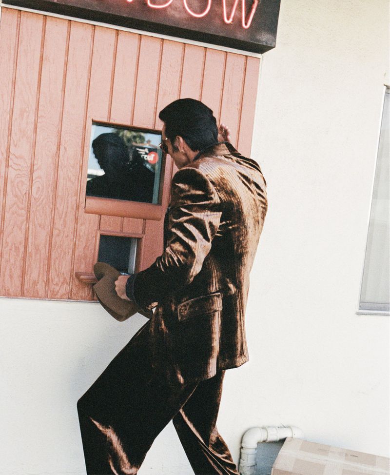 A stylish adult male in a brown suit interacting at an outdoor service window, partially obscured by his hat, with a neon sign overhead.