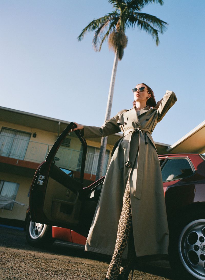 Stylish woman in trench coat opens car door next to a retro car, palm tree, and building on a sunny day in an outdoor fashion shot.