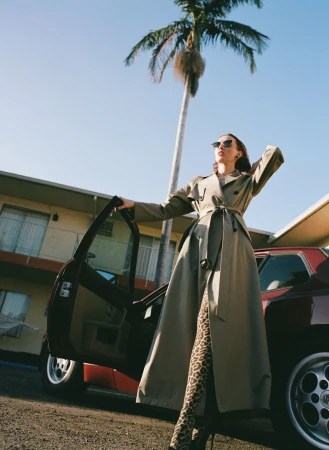 Stylish woman in trench coat opens car door next to a retro car, palm tree, and building on a sunny day in an outdoor fashion shot.
