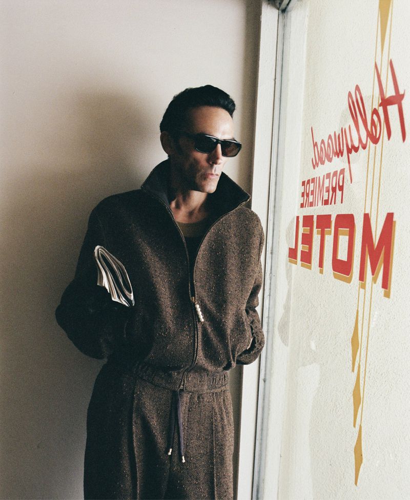 Adult male in brown tracksuit and sunglasses stands next to Hollywood Premiere Motel sign in an indoor setting with neutral light.
