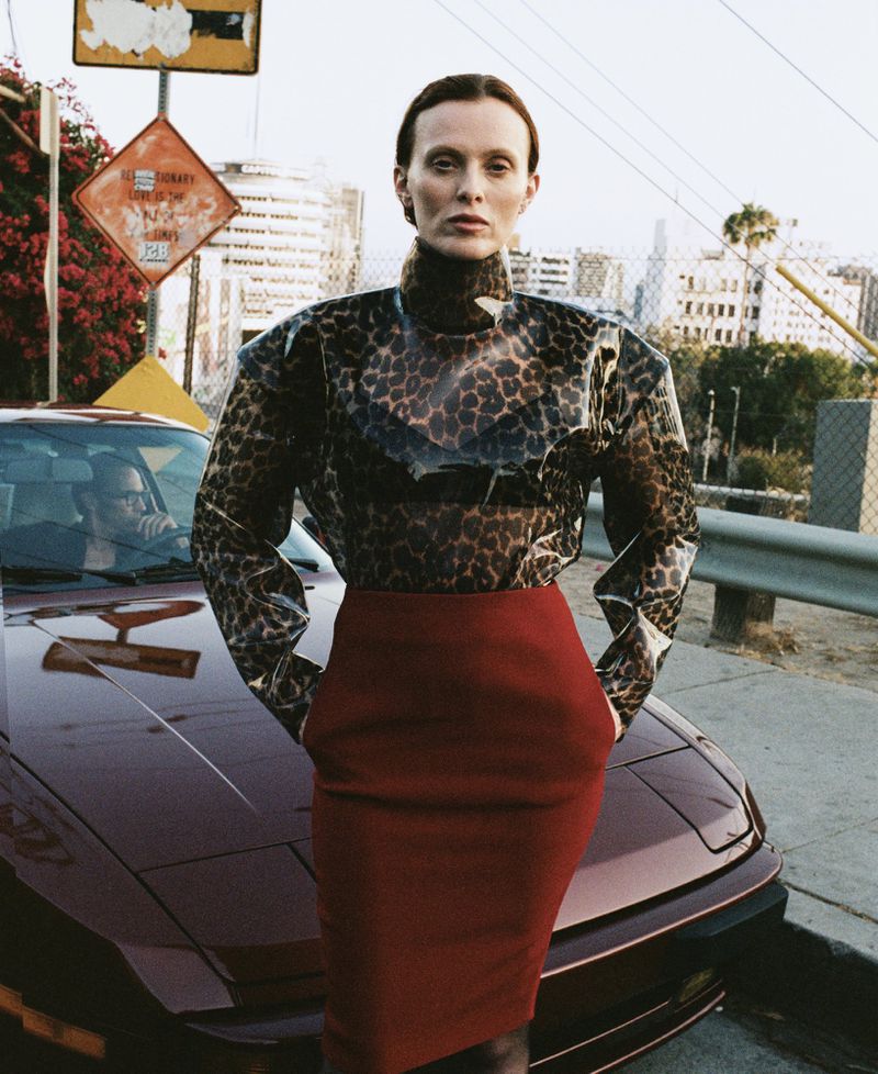 A woman in a leopard-print top and red skirt poses next to a burgundy car on an urban street with buildings in the background.