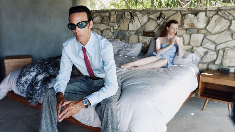 Stylish man and woman in bedroom. Man in suit sits on bed's edge, woman in dress reclines on bed, stone wall behind, natural light.