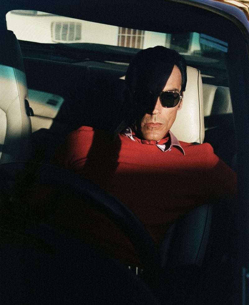 An adult male in sunglasses driving a car, partially obscured by shadow, wearing a red shirt with a white collar, car interior.