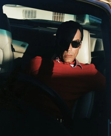 An adult male in sunglasses driving a car, partially obscured by shadow, wearing a red shirt with a white collar, car interior.