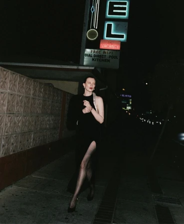 Adult female walking in black dress and heels next to a motel at night under neon signs with dark lighting.