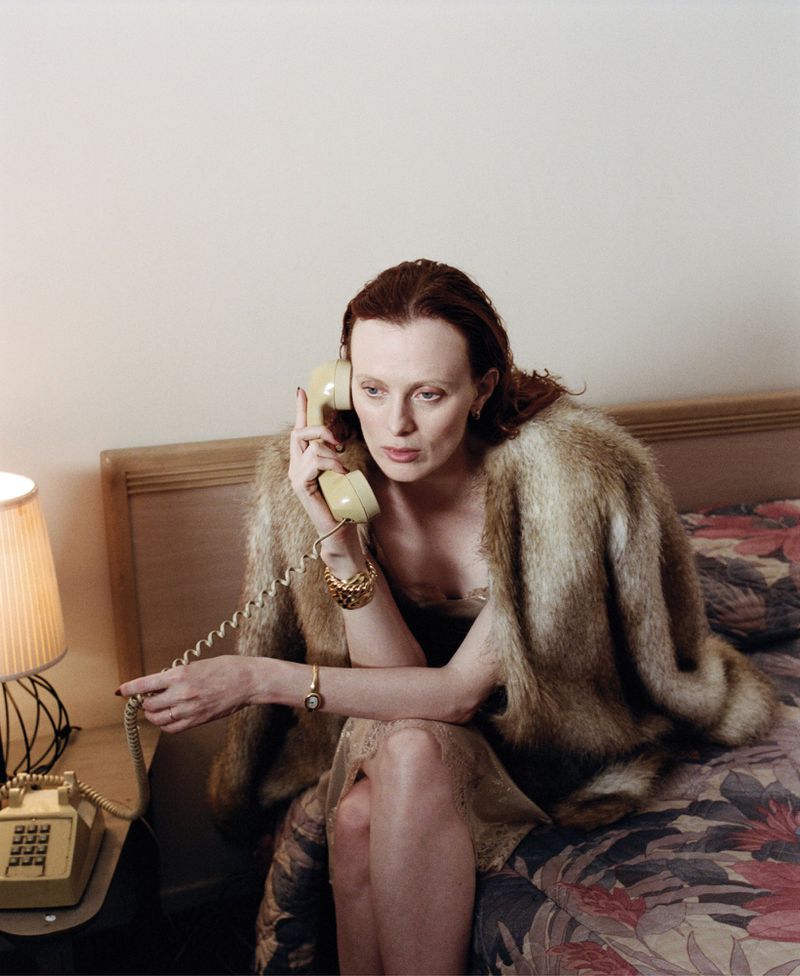 Woman in fur coat sits on bed using vintage rotary phone in indoor shot. She wears gold jewelry and has long auburn hair.