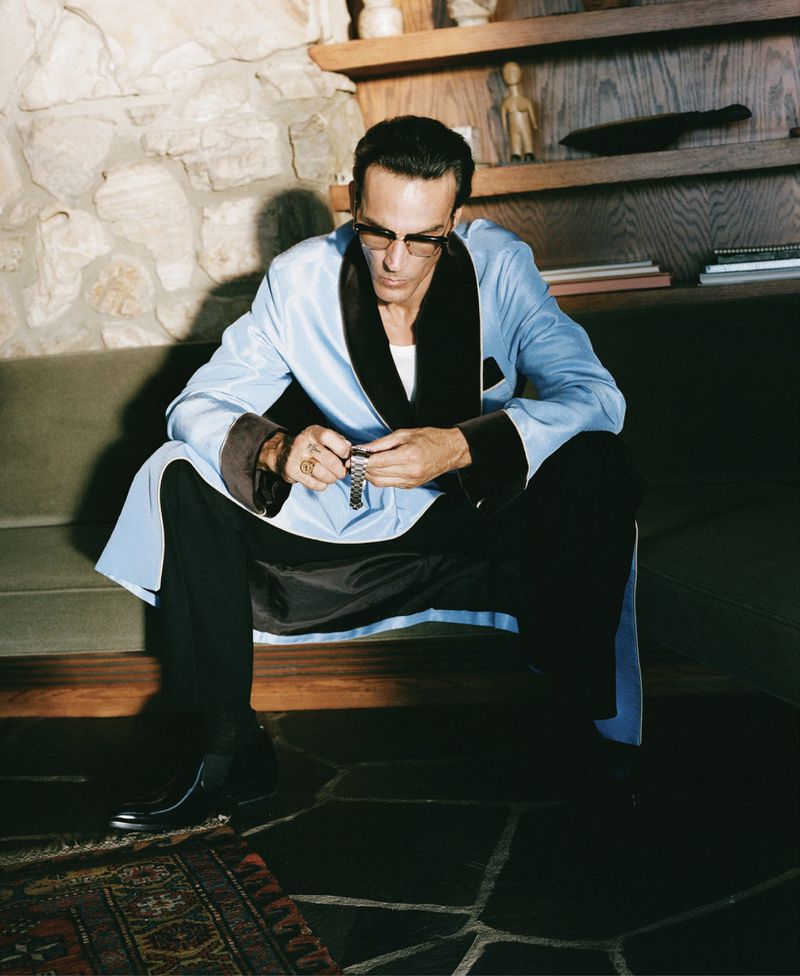 Adult male wearing glasses and a blue robe seated on a stone step inside a house with a stone fireplace and display shelves.