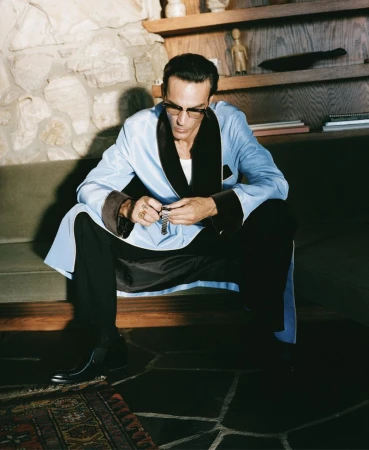 Adult male wearing glasses and a blue robe seated on a stone step inside a house with a stone fireplace and display shelves.