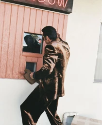 Model Scott Barnhill in a shiny brown suit holding a hat while leaning towards a wooden service window beneath a pink neon sign.