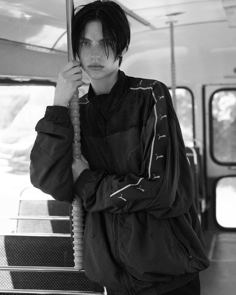 Monochrome portrait of a person with dark hair wearing a Puma tracksuit jacket inside of a bus with a neutral expression.