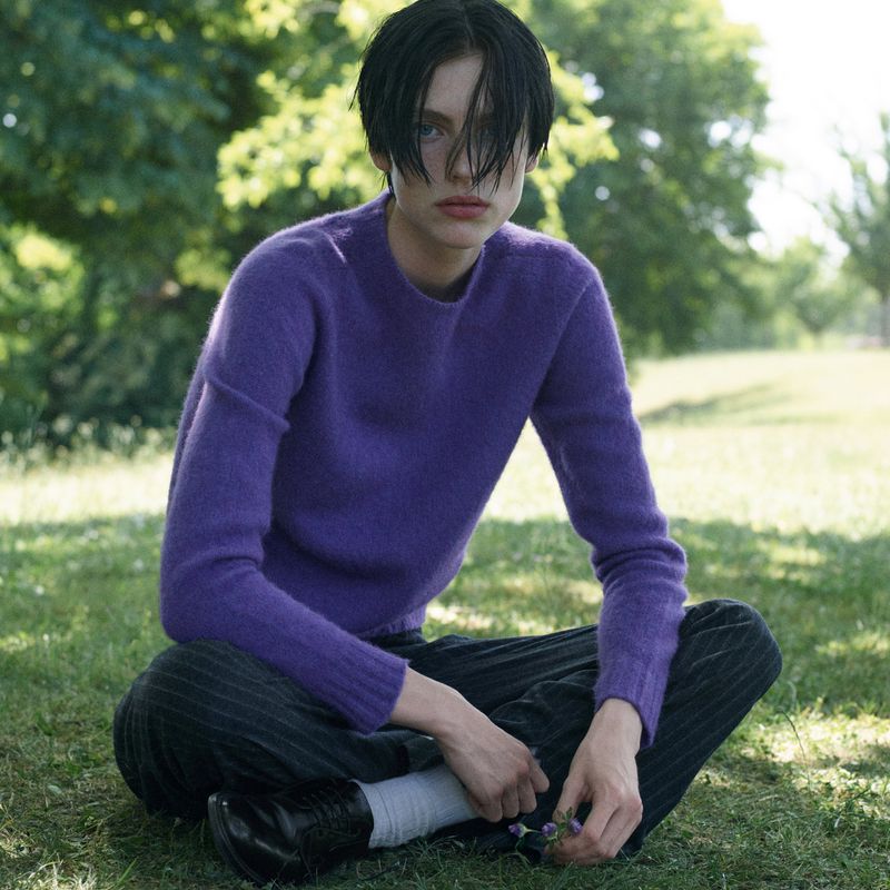 Woman in purple sweater sits cross-legged on grass in a park-like setting, with soft focus on face and upper torso, casual outdoor fashion.