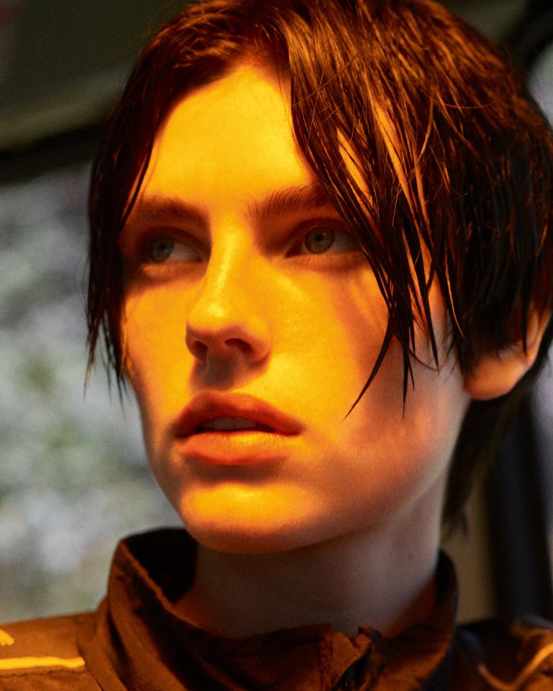 Close-up portrait of a young adult female with short brown hair, wearing a dark jacket, lit with golden-yellow light in an outdoor setting.